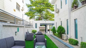 Un patio con mobiliario exterior, un árbol y un edificio de fachada blanca.