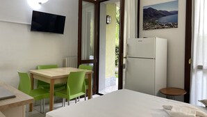 Habitación de hotel con cama, escritorio con una computadora portátil, mesa de comedor con sillas, televisor montado en la pared, refrigerador y ventana con vista a un lago y montañas.