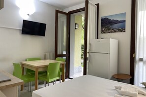 A hotel room with a bed, a desk with a laptop, a dining table with chairs, a TV mounted on the wall, a refrigerator, and a window with a view of a lake and mountains. - Mobilehome am Luganersee (Porlezza)