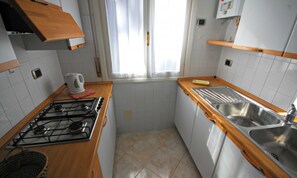 A small kitchen with a gas stove, a sink, and a window with curtains. - Cozy Villa With Patio and Garden (Lignano Sabbiadoro)