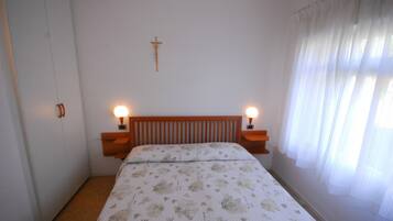 A bedroom with a wooden headboard, two bedside lamps, a bed with floral bedding, a cross on the wall, and a window with sheer curtains.