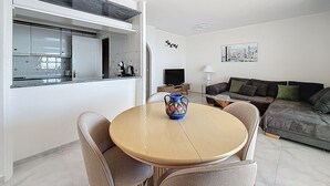 A modern living room with a round wooden table, a sofa, and a kitchen area with a window. - BELLA PANORAMICA Roses / Roses - INTERimmo (Rosas / Roses)