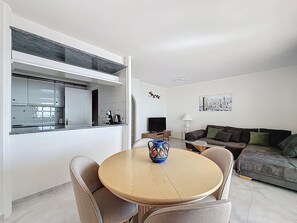 A modern living room with a round wooden table, a sofa, and a kitchen area with a window. - BELLA PANORAMICA Roses / Roses - INTERimmo (Rosas / Roses)