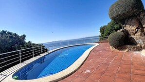 A curved swimming pool with a clear blue water surface, surrounded by a red-tiled deck and a metal railing. - BELLA PANORAMICA Roses / Roses - INTERimmo (Rosas / Roses)