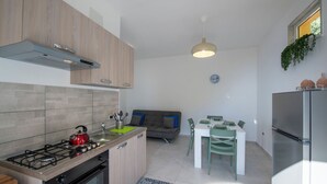 A modern kitchen with a gas stove, oven, and a built-in refrigerator. There is a dining area with a table and chairs, and a sofa in the background. - VivimiHome - Casa Angelina (Conca dei Marini)