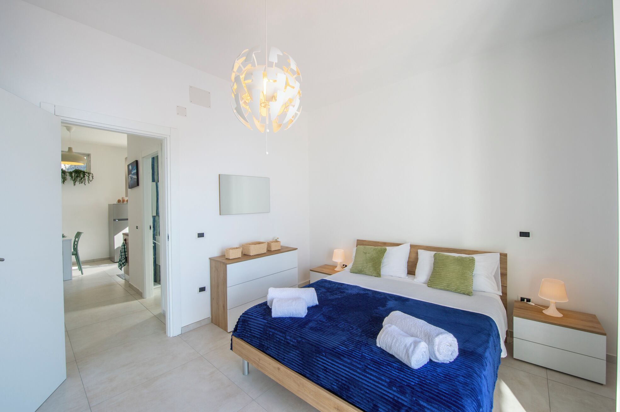 Une chambre moderne avec un grand lit, des tables de chevet, un luminaire suspendu et une vue sur la pièce adjacente.