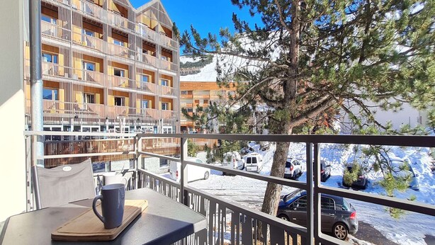 Un balcón con una mesa y una silla, con vista a una calle nevada y un edificio de varios pisos con balcones.