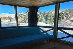 A room with a blue sofa, large windows, and a view of a snowy landscape with trees and a building. - Ski Base for 4 near La Foux Lifts (Allos)