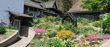Un poble europeu tradicional amb edificis de pedra, un camí empedrat i una varietat de flors de colors al jardí.