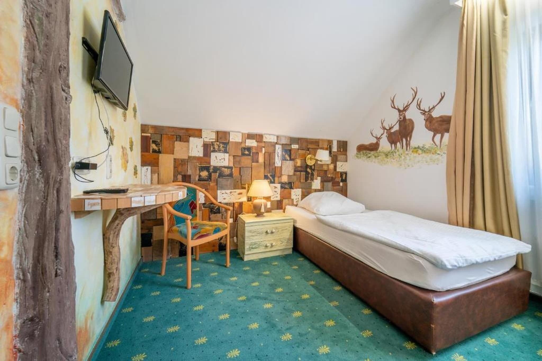 Una habitación con un escritorio de madera, una silla, una cama, una mesita de noche, una lámpara y un mural de un ciervo en la pared.