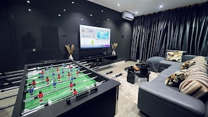 Ein modernes Wohnzimmer mit einem Kicker, einem Flachbildfernseher, dunklen Vorhängen und einer Couch mit gemusterten Kissen.