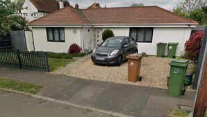 A single-story house with a car parked in the driveway, a fenced yard, and two green waste bins. - Step-Free Bungalow w/ Garden & Parking, Near LGW (Wallington, Surrey)