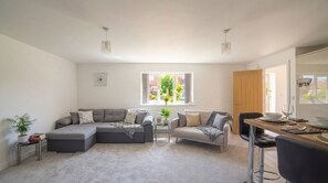 A modern living room with a sectional sofa, a small table with a plant, and a dining area with a bar stool. - 2-Bed Wallington, Sleeps 6, Parking, Near LGW (Wallington, Surrey)