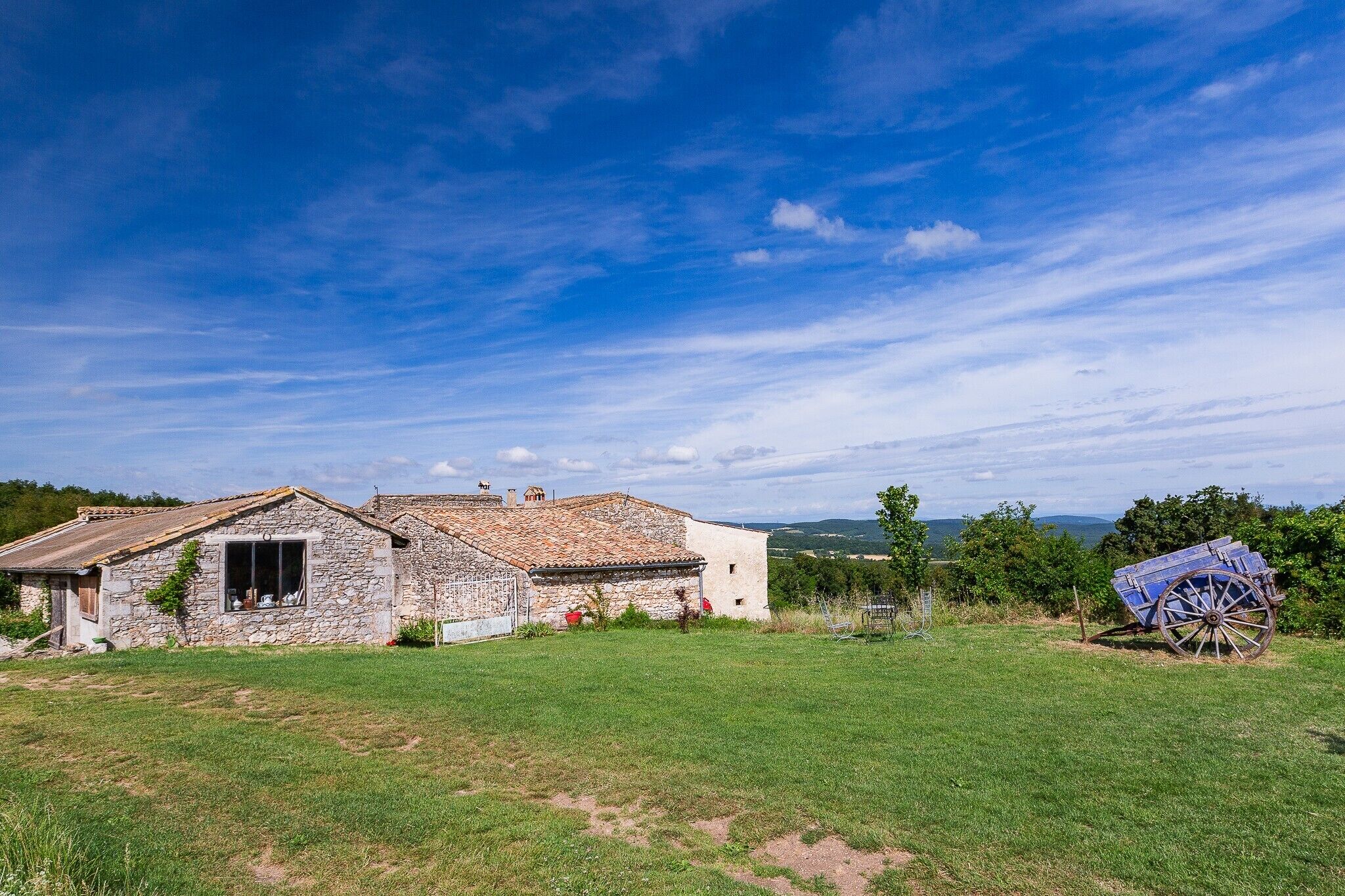 Une ferme en pierre avec un toit de tuiles, un champ verdoyant et un chariot au premier plan.