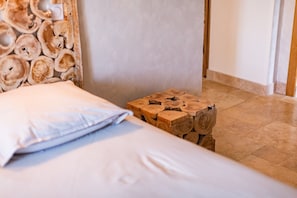 Uma cama com lençol branco e travesseiro, um banco de tronco de madeira e uma parede com design em ripas de madeira.
