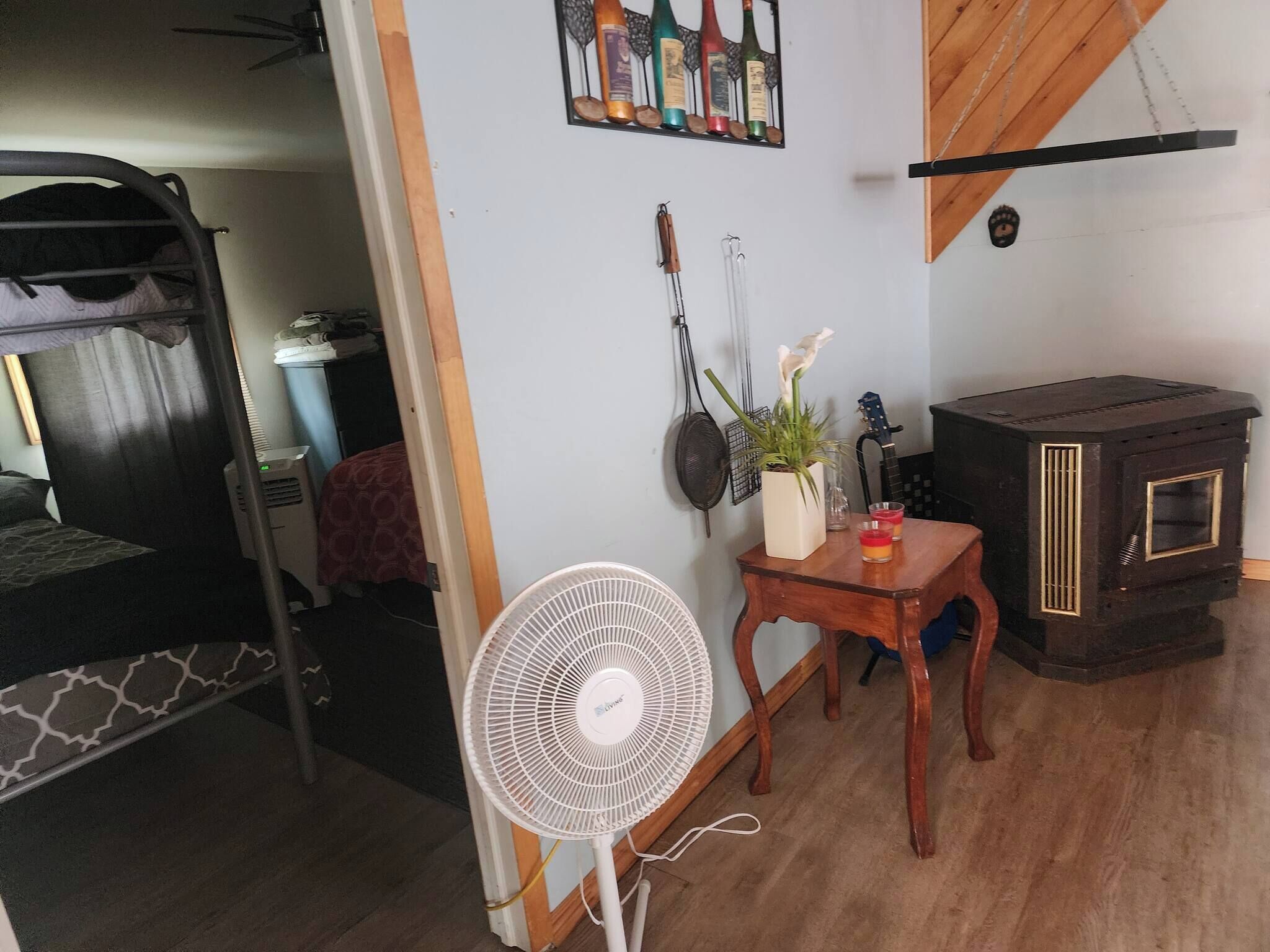 A room with a bunk bed, a fan, a table with a plant, a guitar, and a wood-burning stove.