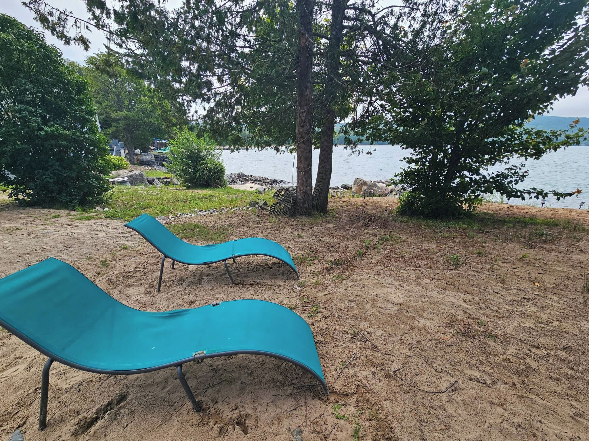 Two blue lounge chairs on a sandy beach with a view of the water and trees in the background.