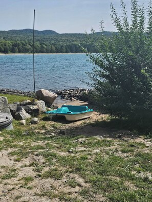 A boat on a sandy shore near a body of water with a forested hill in the background. - Lake Superior Waterffront Main Unit of Duplex Sleeps 8 ppl (Unorganized North Algoma)