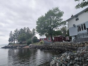 A waterfront property with a house adjacent to a rocky shoreline, a stone wall, and a calm body of water. - Lake Superior Waterffront Main Unit of Duplex Sleeps 8 ppl (Unorganized North Algoma)