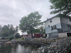 A waterfront property with a stone retaining wall, a large tree, and a two-story building with a balcony. - Lake Superior Waterffront Main Unit of Duplex Sleeps 8 ppl (Unorganized North Algoma)