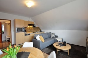 A compact living space with a kitchenette, a grey sofa with cushions, a small dining table, and a round coffee table. - Haus Pithan - House Pithan in Werdum - Holiday home with garden, WLAN and 4 (Werdum)