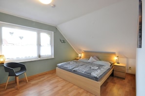 A bedroom with a sloped ceiling, a bed with striped bedding, a nightstand with a lamp, a chair, and a window with a view of buildings. - Haus Pithan - House Pithan in Werdum - Holiday home with garden, WLAN and 4 (Werdum)