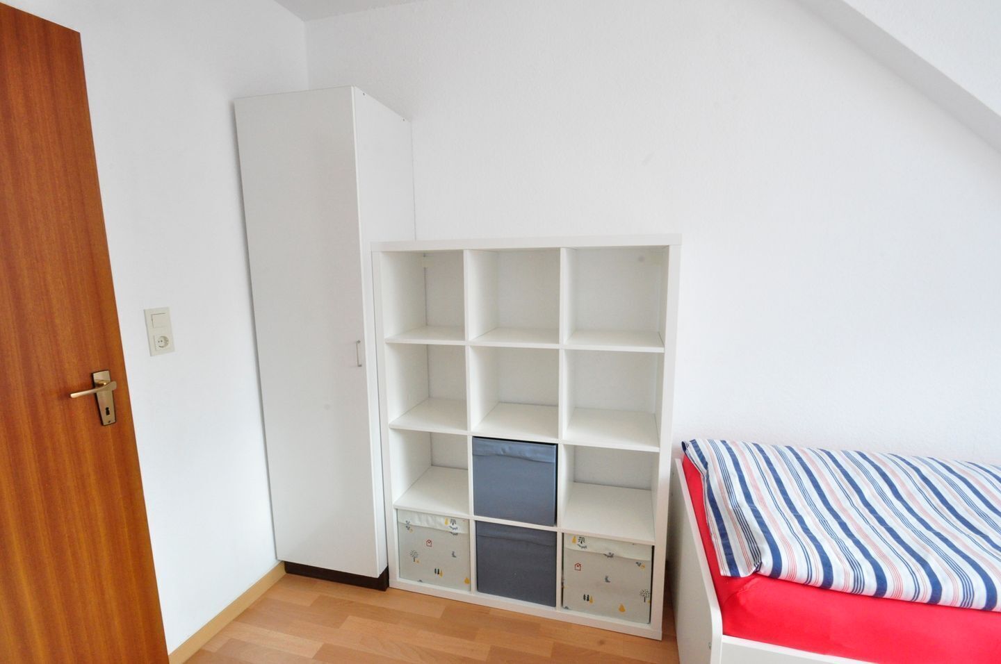 A room with a wooden door, a white wardrobe, a bed with striped bedding, and a white shelving unit with storage boxes.