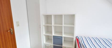 Una habitación con puerta de madera, un armario blanco, una cama con ropa de cama a rayas y un estante blanco con cajas de almacenamiento.