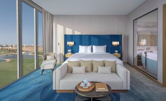 A modern hotel room with a large bed, a sofa, a dining table, and a view of a golf course.