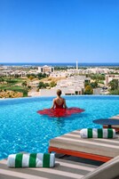 A pool with a person in a red swimsuit, lounge chairs, and a view of a coastal town.