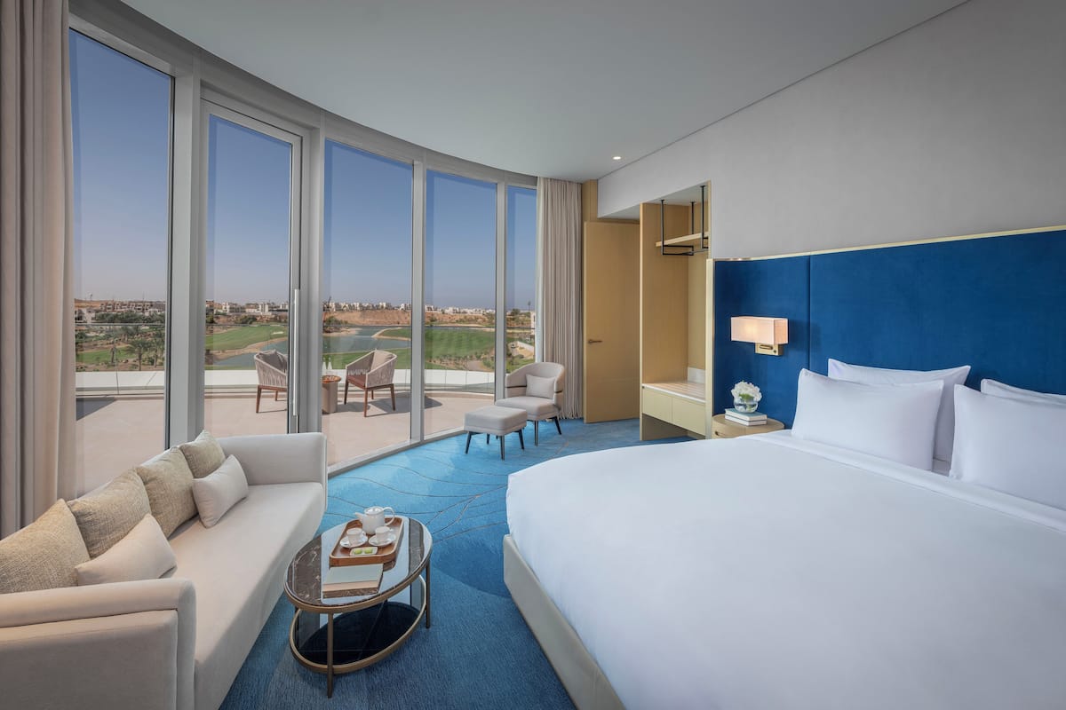 Une chambre d’hôtel avec un grand lit, un canapé, une petite table avec un plateau, une chaise et une vue sur un terrain de golf.
