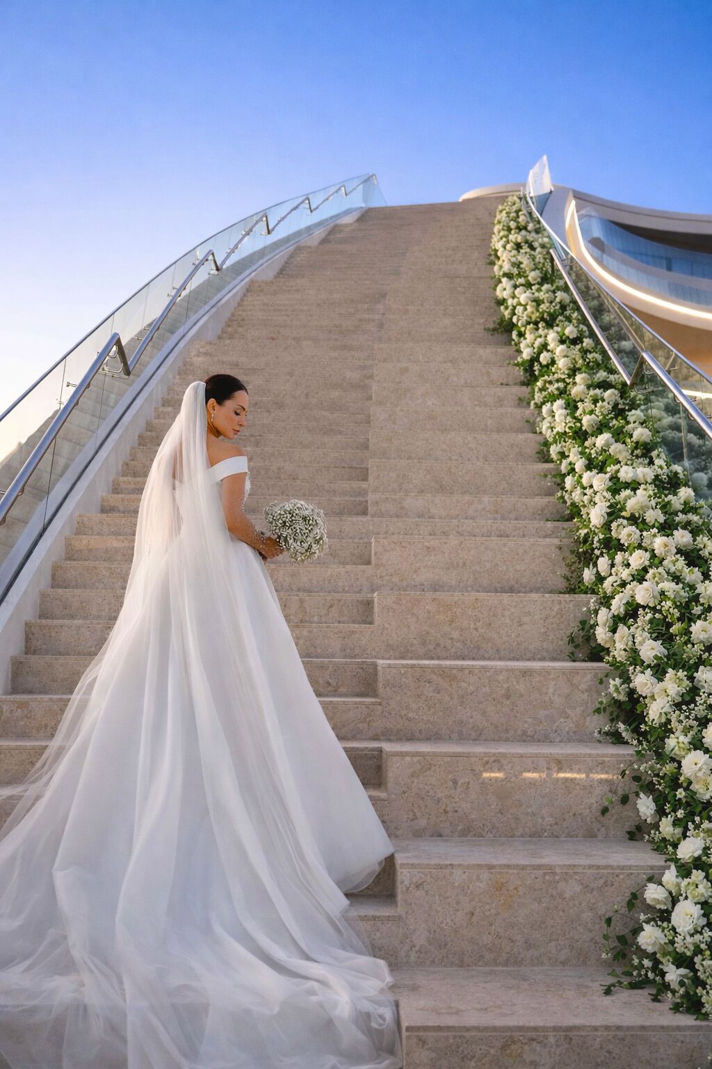 A bride in a white wedding dress descending a staircase with a floral arrangement on the right.
