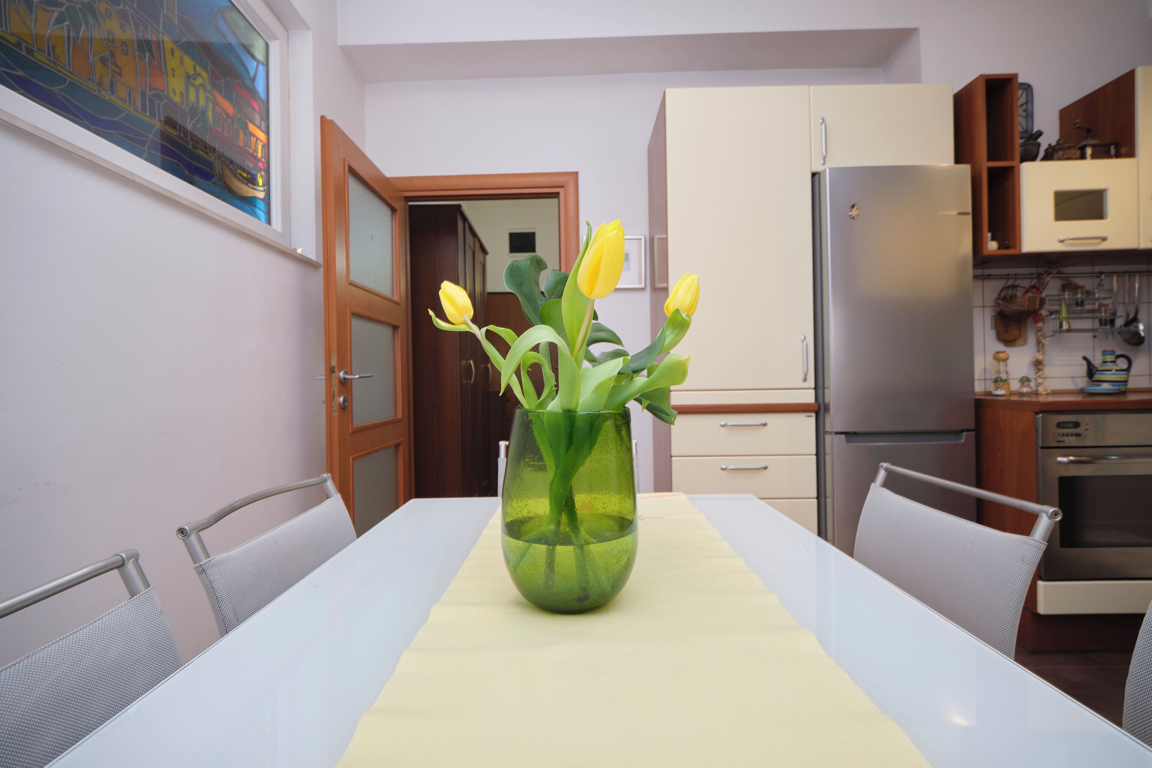A dining table with a vase of yellow flowers, a white tablecloth, and a view of a kitchen with cabinets and appliances.
