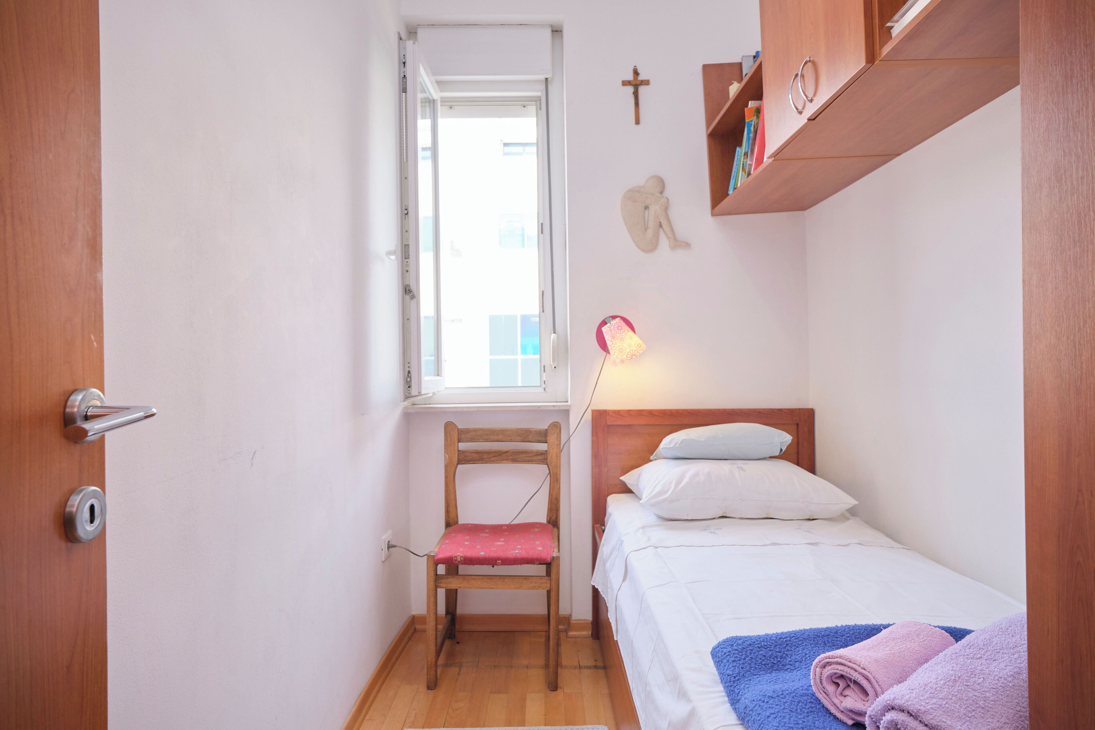 A small, well-lit room with a single bed, a chair, a window, and a shelf with books.