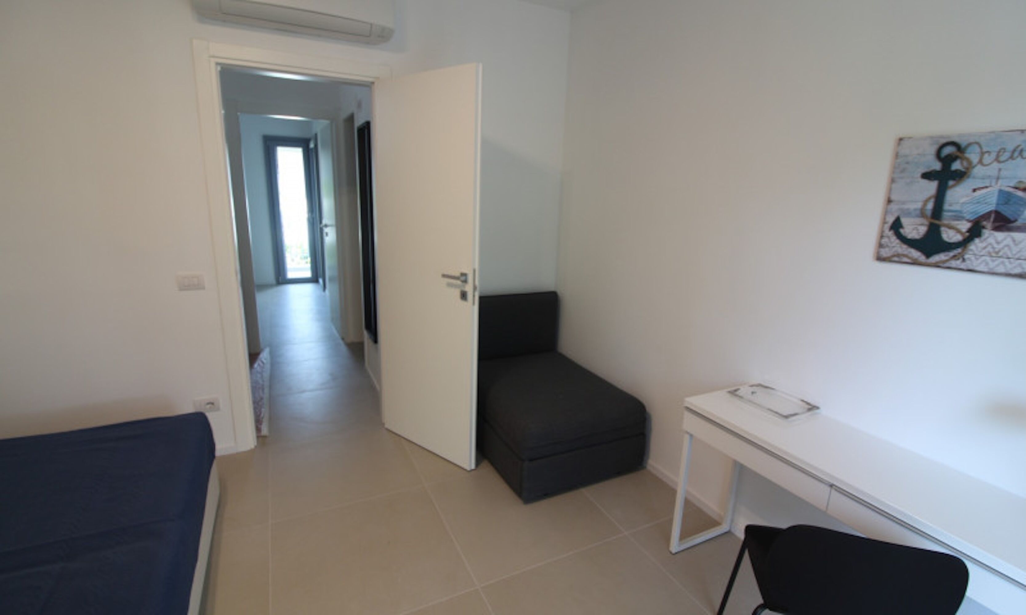 A room with a bed, a desk, and a chair. There is a door leading to another room.