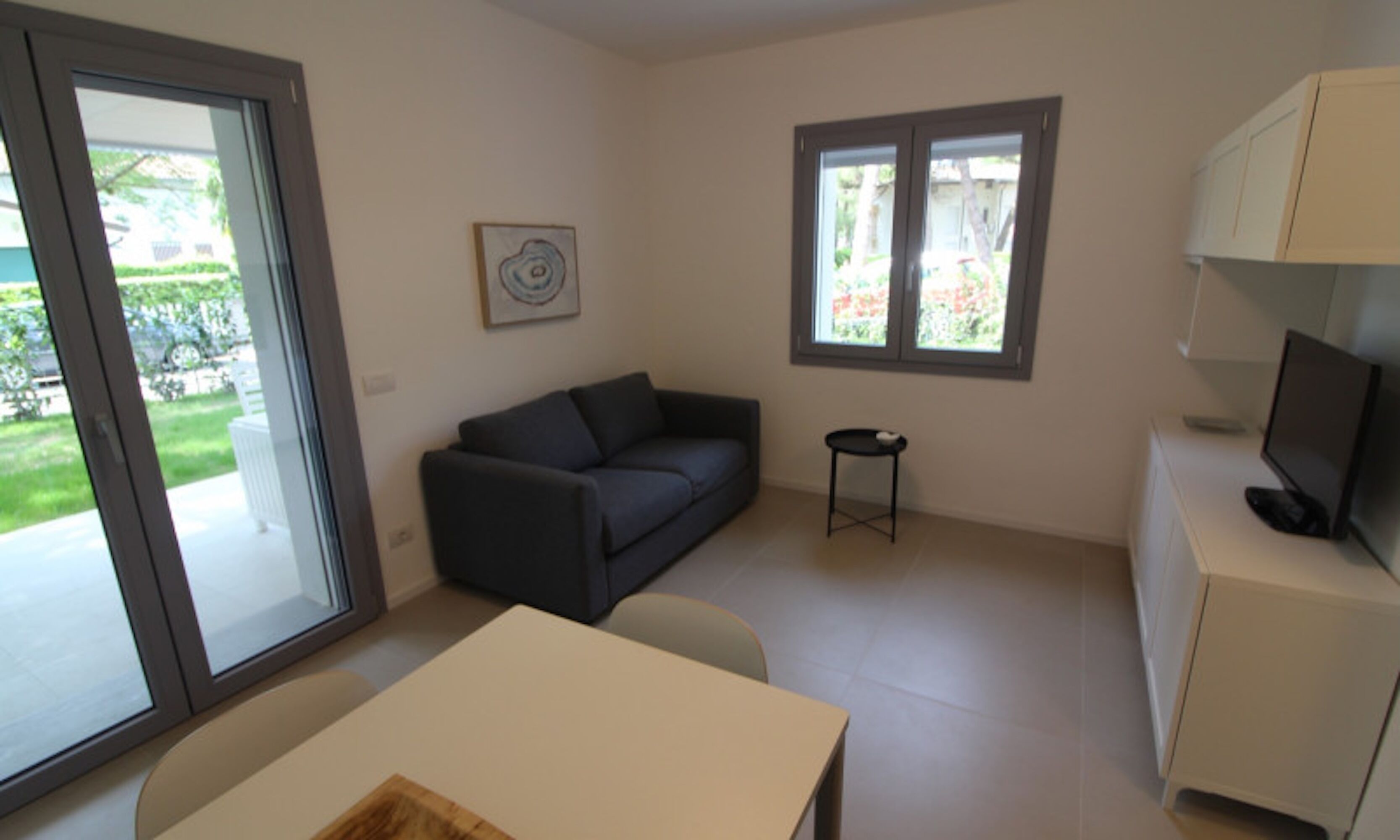 A modern living room with a sofa, a small table, a TV, and a window with a view of greenery.