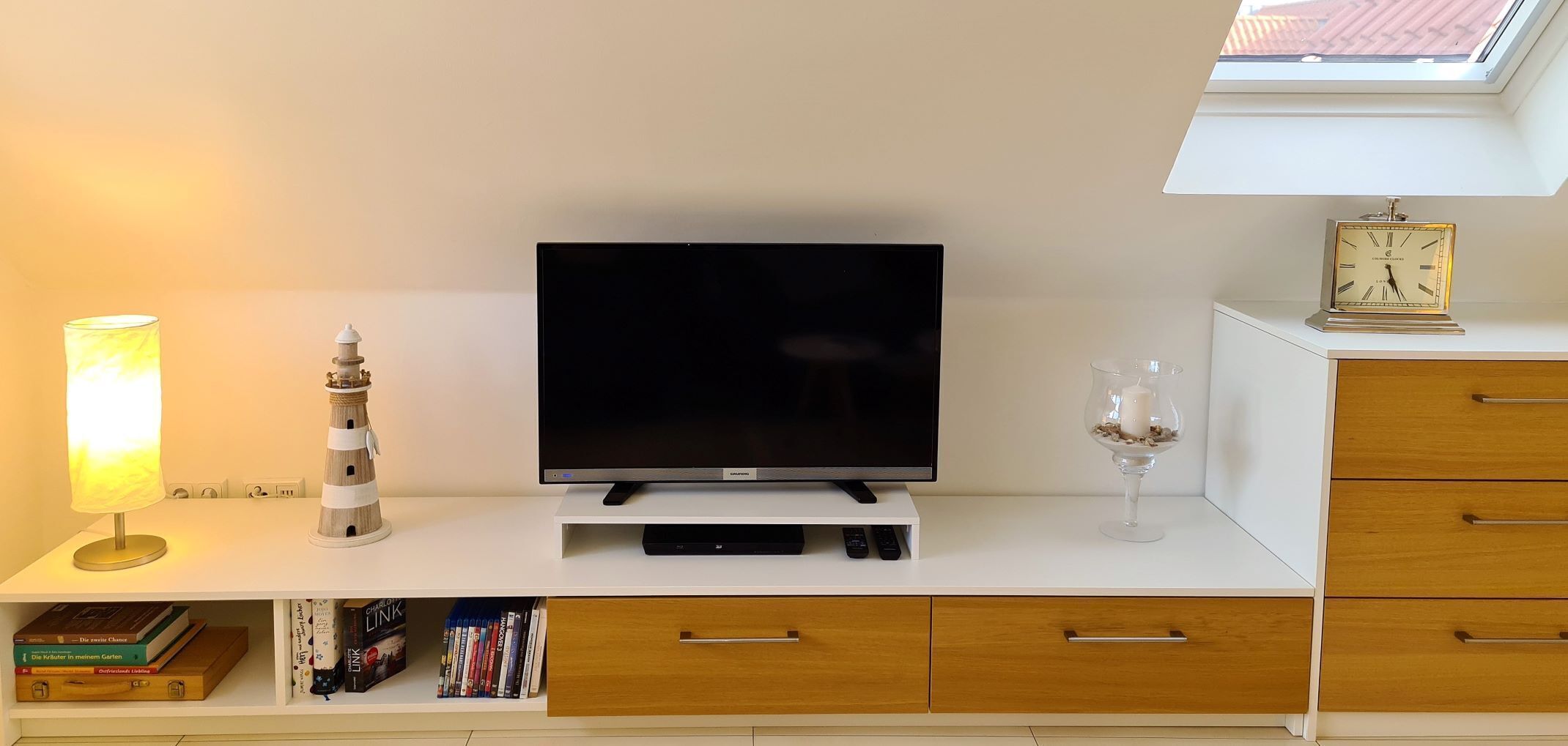 Moderni olohuone, jossa on valkoinen, litteänäyttöinen televisio, pieni kirjahylly kirjoineen, koristeellinen majakka, valaistu pöytälamppu, kynttilä lasissa ja seinällä oleva kello.
