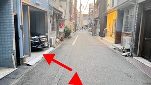 Una calle urbana estrecha flanqueada por edificios, un coche aparcado y flechas rojas indicando la dirección en la calzada.