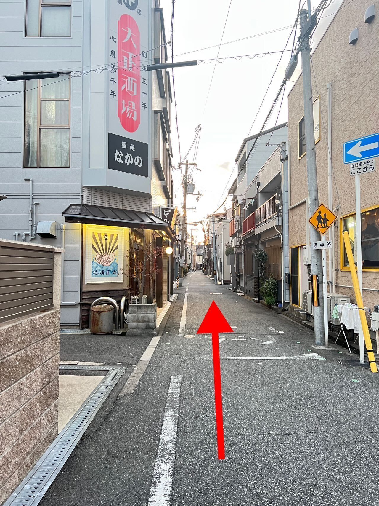 A narrow urban street with buildings on either side, a visible storefront with a sign, and a directional arrow on the road.