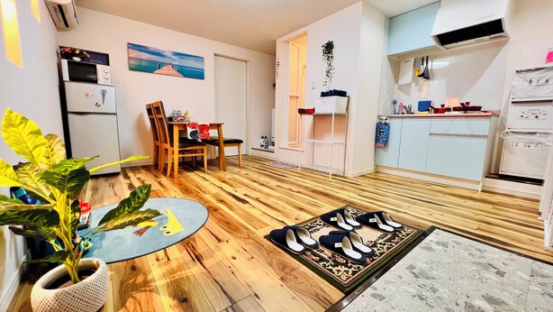 Un espacio habitable compacto con piso de madera, un comedor con sillas, una mesita, una planta en maceta, una zona de cocina con armarios y electrodomésticos, y un aire acondicionado instalado en la pared.