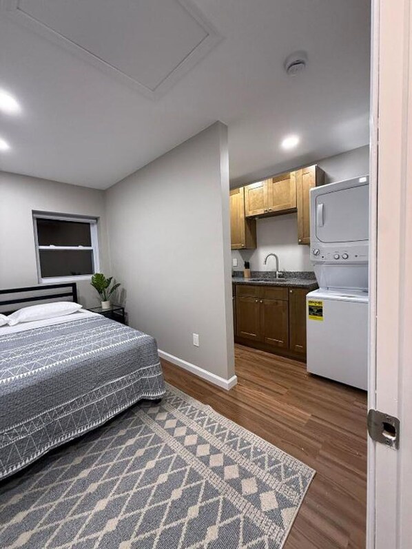 A compact bedroom with a bed and a window, a kitchenette with wooden cabinets and a washing machine, and a patterned rug on the floor.