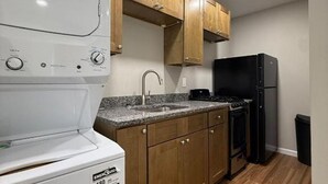 A compact kitchen with wooden cabinets, a white washing machine, a black refrigerator, and a granite countertop.