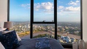 Un dormitorio con un ventanal grande que ofrece vistas panorámicas de la ciudad, una cama con ropa de cama estampada con motivos florales y una lámpara de noche.