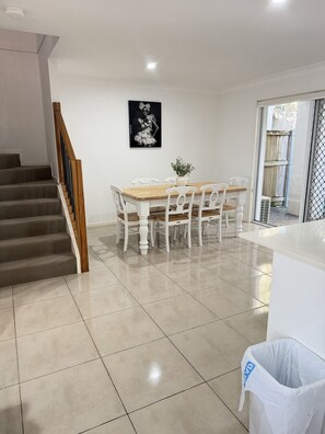 A dining area with a wooden table and white chairs, a staircase with wooden railings, and a white trash bin. - Syldah Stay Mango Hills (Mango Hill)
