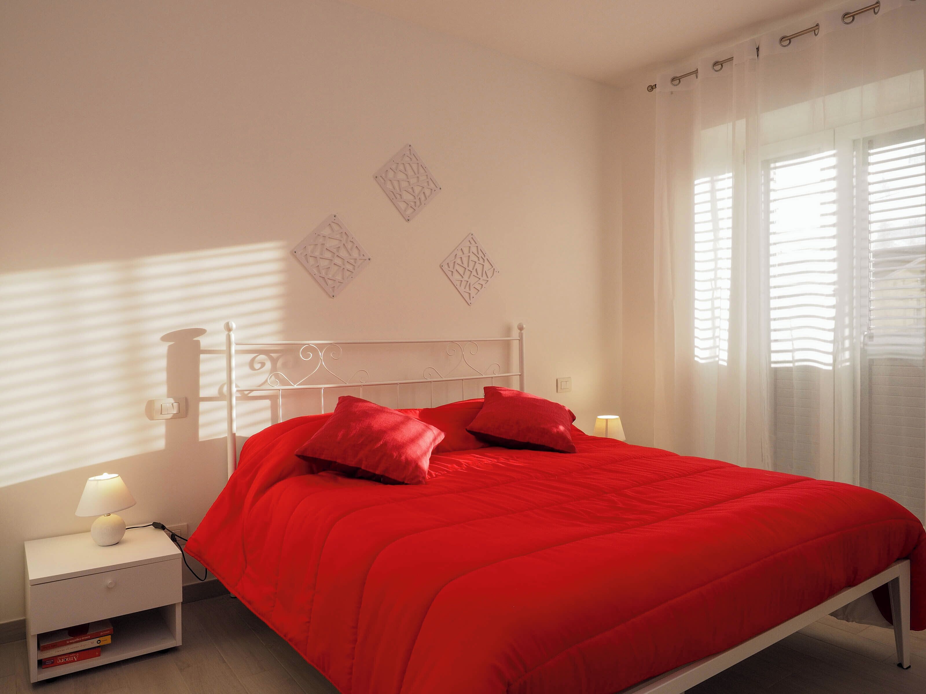 A bed with a red bedspread and two pillows, a bedside table with a lamp, and a window with blinds.