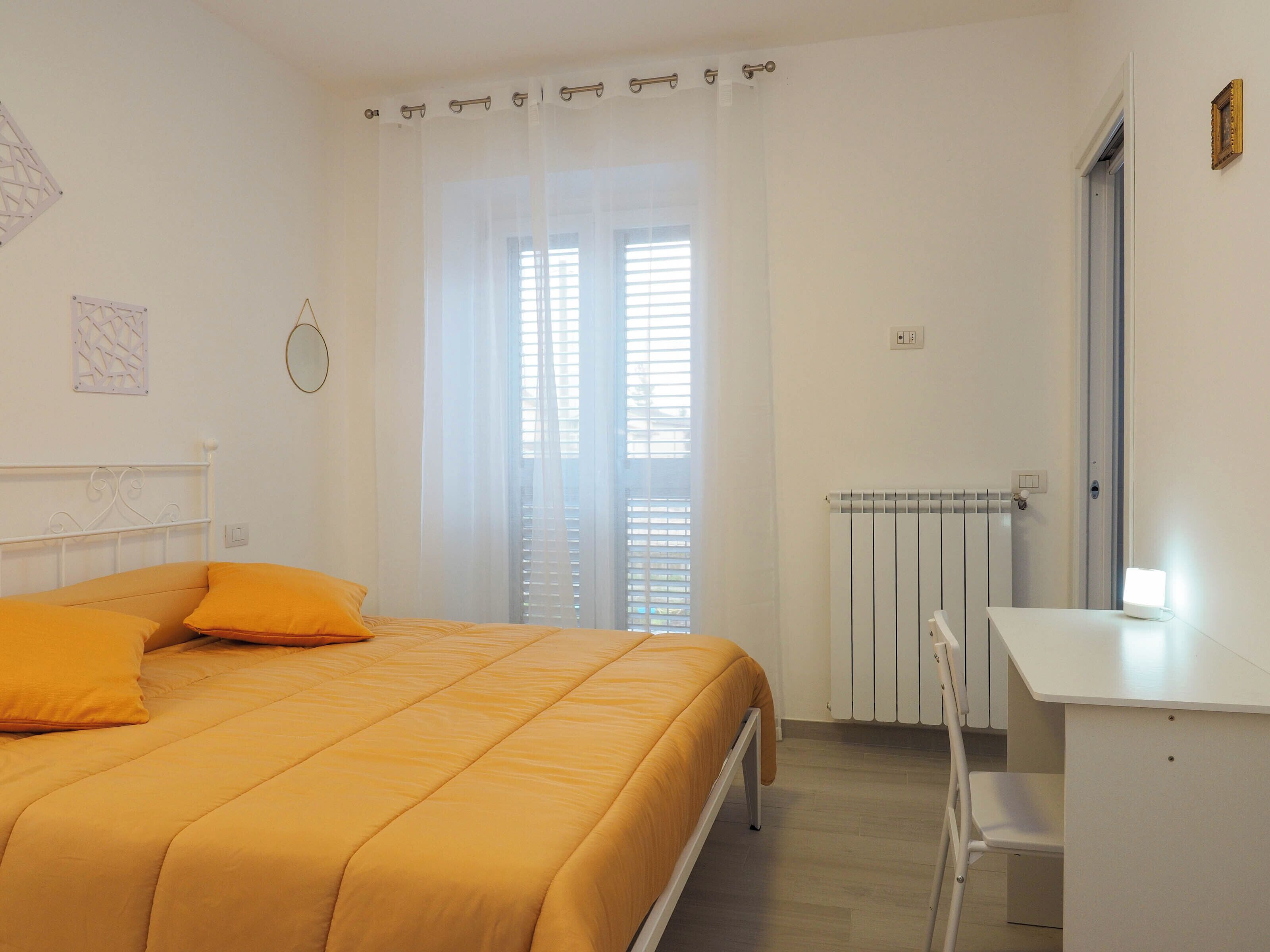 A bedroom with a bed, a desk, a radiator, and a window with blinds.