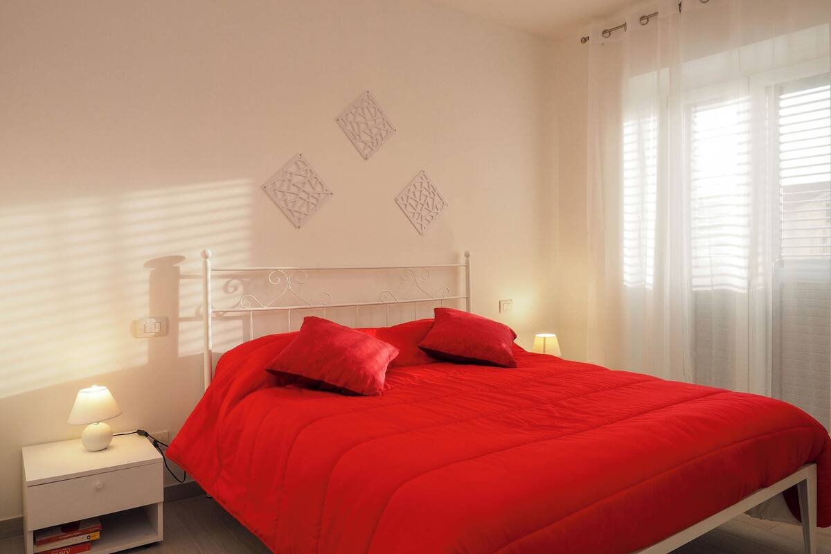 A bedroom with a white metal bed frame, a red bedspread, and two red pillows. There is a bedside table with a lamp and a book. The room has white walls and a window with sheer curtains.