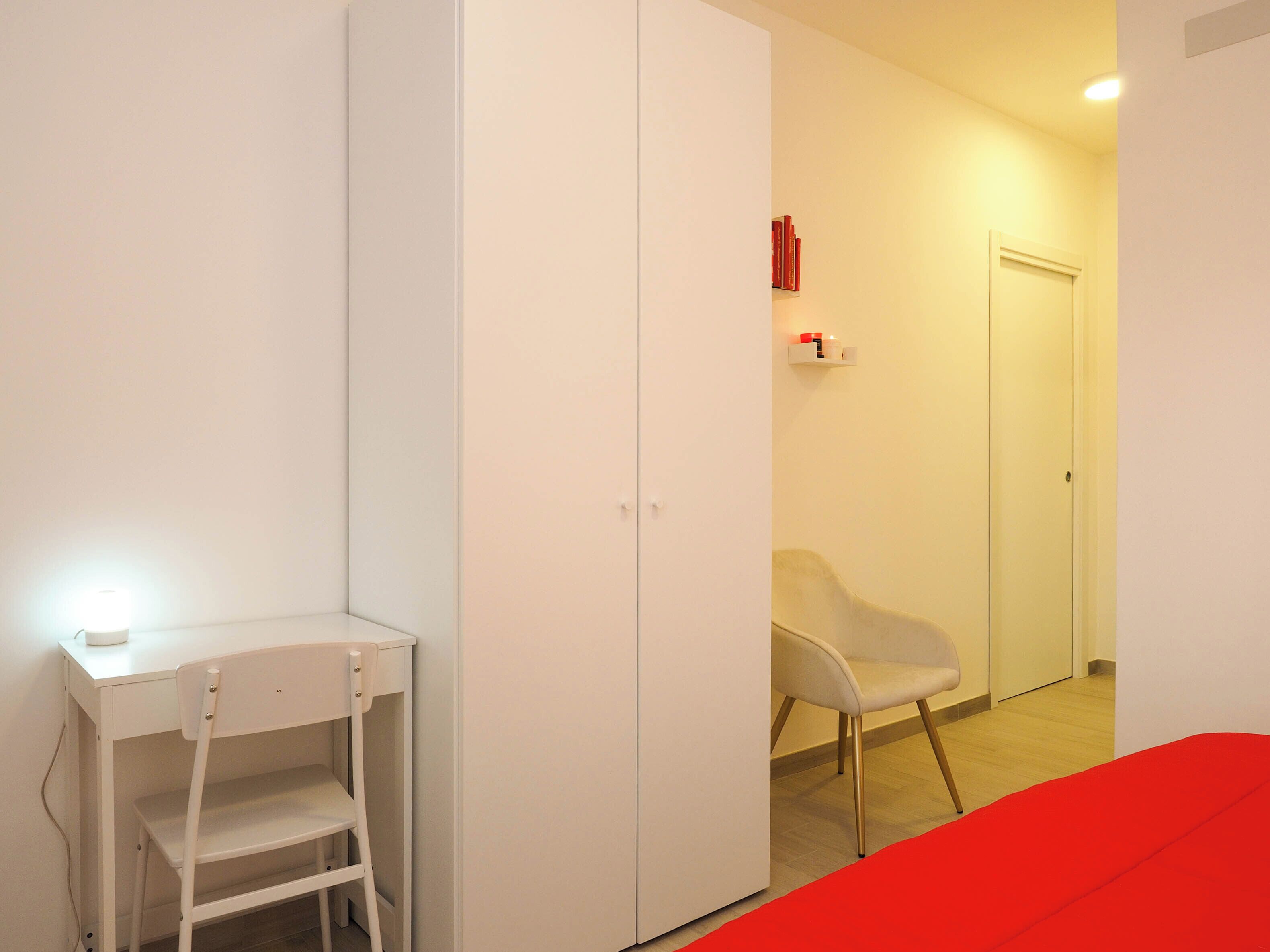 A room with a white desk, a chair, a wardrobe, and a door.