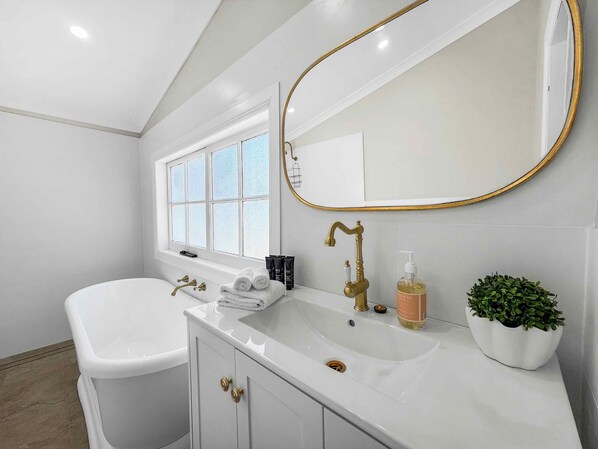 Un baño moderno con una bañera blanca, un espejo grande con marco dorado y una pequeña planta en el mueble de tocador.