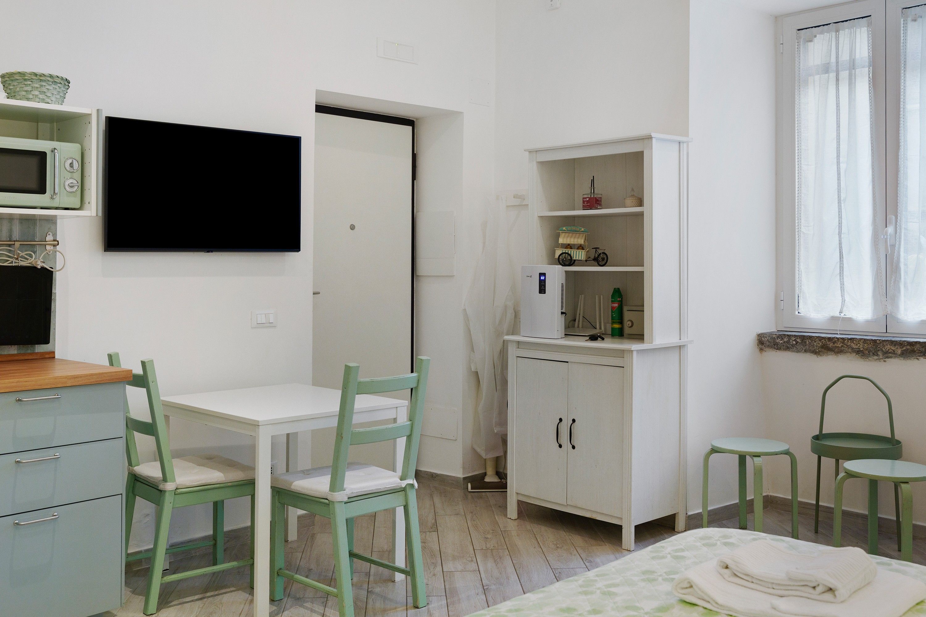 A room with a white table, two chairs, a microwave, a cabinet with shelves, and a window with curtains.
